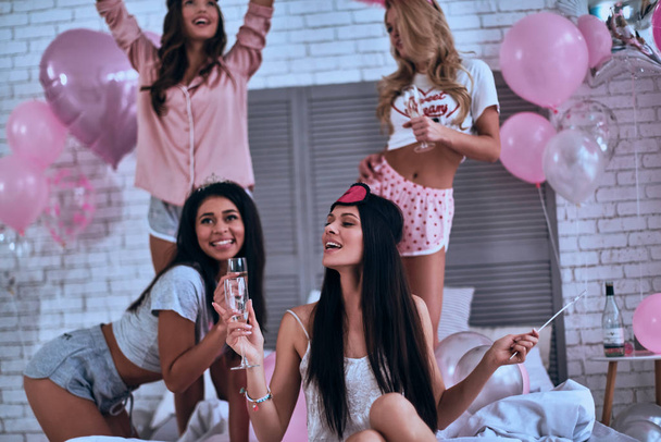 Four attractive young smiling women in pajamas drinking champagne while having a bachelorette party in bedroom - Φωτογραφία, εικόνα