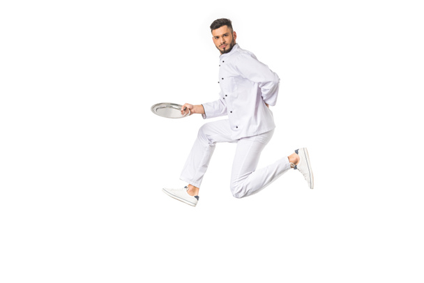 young male chef holding plate while jumping and looking at camera isolated on white - Photo, Image