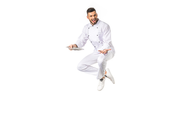 happy young male chef with plate jumping and smiling at camera isolated on white - Photo, Image