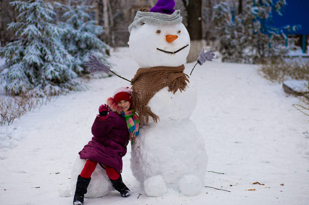 Küçük bir neşeli kız büyük gerçek komik kardan adam oturur. Sevimli küçük bir kız kış Park, eğlenir kış - Fotoğraf, Görsel