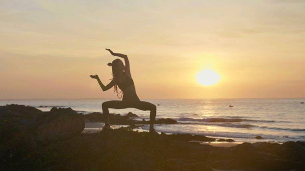 Siluettijoogaharjoitukset auringonlaskun aikaan. Yong nainen tekee jooga liikuntaa rannalla
 - Valokuva, kuva