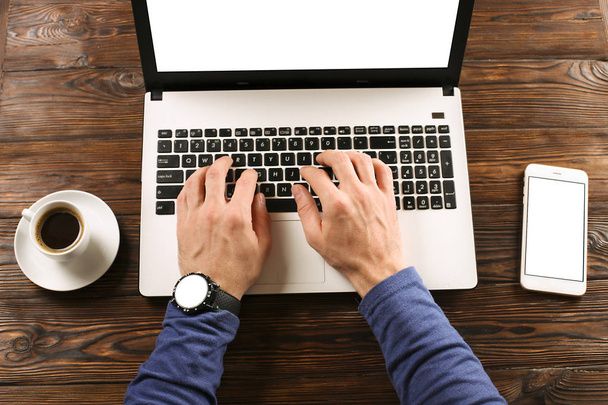Niedbale ubrany student / blogger / pisarz / man działa na komputerze przenośnym, wpisując na klawiaturze, pisanie blogu, picie, kawiarnia / bar, za pomocą smartfonu. Widok z góry z biurka roboczego. - Zdjęcie, obraz