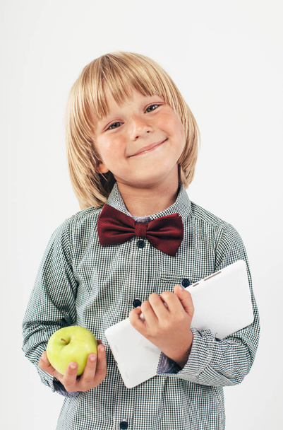 Smiling School boy in shirt with red bow tie, holding tablet computer and green apple in white background - Photo, Image