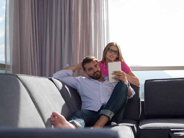 Молода пара розслабляється вдома за допомогою планшетних комп'ютерів, які читають у вітальні на дивані
. - Фото, зображення