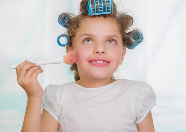 Little girl using a brush to make up while wearing hair-rollers and bathrobe in a blurred background - Fotografie, Obrázek