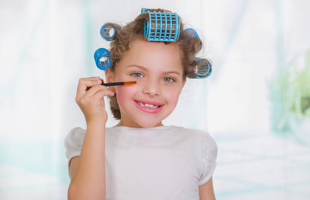 Little girl using a eye mascara in her eye while wearing hair-rollers and bathrobe in a blurred background - Foto, afbeelding