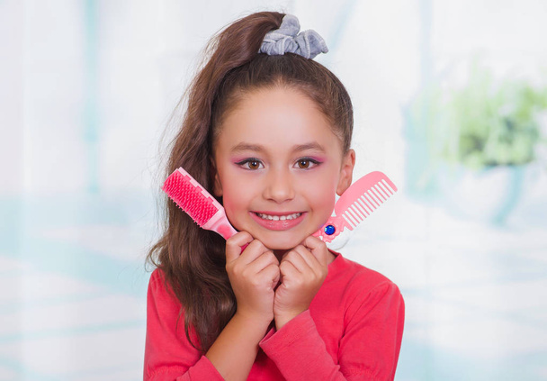 Little beautiful girl wearing a red blouse and holding a hair brush and comb in her hand, in a blurred background - Photo, image