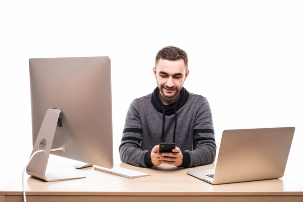 Pohledný podnikatel sedí za svým stolem s laptopem a pc a psaní na mobilním telefonu izolovaných na bílém pozadí - Fotografie, Obrázek