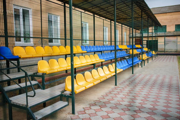 Пустые синие и желтые спортивные сиденья большой трибуны на заднем дворе школы на стадионе
 - Фото, изображение