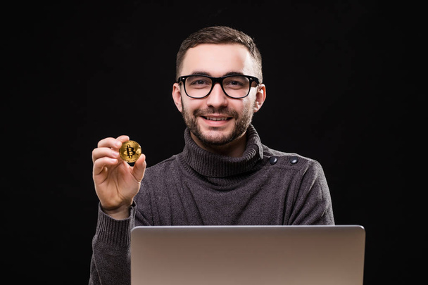 Portrait of a happy businessman showing bitcoin while sitting at desk with laptop computer isolated over black background - Zdjęcie, obraz