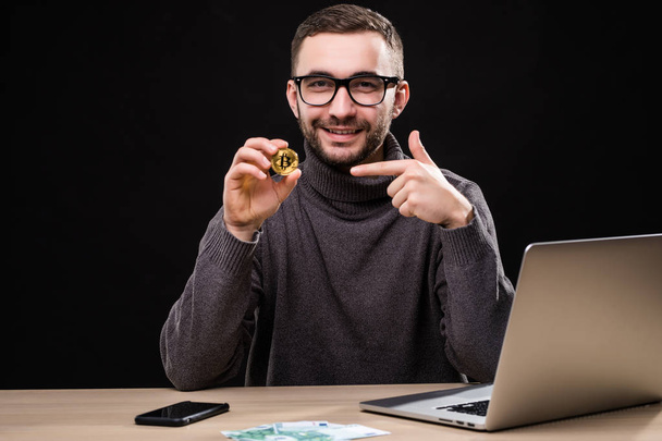 Retrato de um empresário feliz apontado em bitcoin enquanto sentado na mesa com computador portátil isolado sobre fundo preto
 - Foto, Imagem
