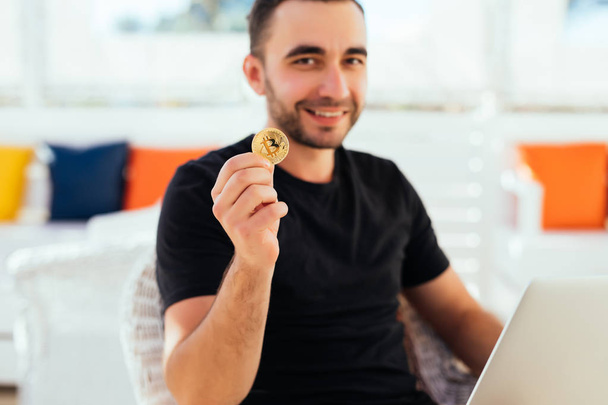 Young handsome man with laptop pointed bitcoin on camera sitting on deck at beack cafe on summer vocation. Man check bitcoin value on summer vocation. - Foto, imagen