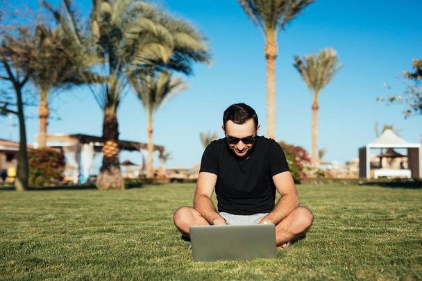 Красивий чоловік сидить на траві і використовує ноутбук, ганяє або читає інтернет-новини на фоні долонь. Літнє покликання
. - Фото, зображення