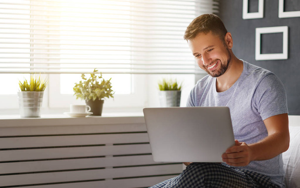 glücklicher junger Mann mit Laptop und Tasse Kaffee im Bett - Foto, Bild