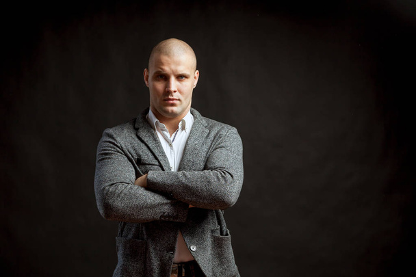 A young bald man in a white shirt, gray suit confidently looks at the camera and holds his arms crossed on his chest on a black isolated background - Photo, Image