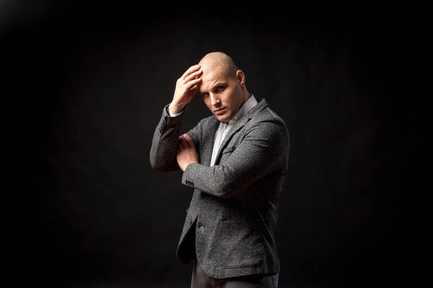 A young bald man in a white shirt and gray suit is upset by a serious problem in business, at work on a black isolated background. A man has a headache - Foto, afbeelding