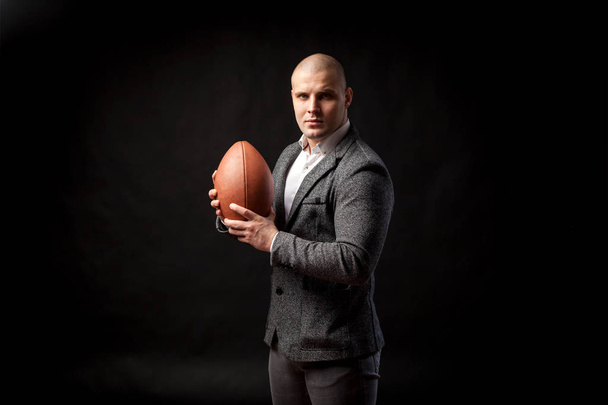 A young bald man in a white shirt, gray suit seriously looks into the camera and keeps a rugby ball on a black isolated background - Photo, Image