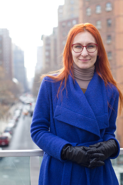 Mavi ceket ve büyük şehrin arka plan üzerinde durup gözlüklü şirin Kızıl saçlı kız - Fotoğraf, Görsel