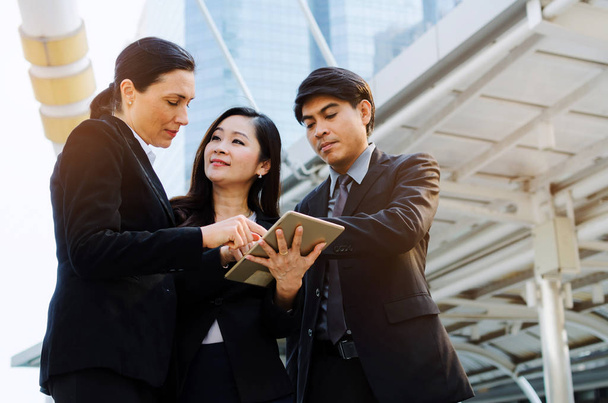 group of business people in suit talking and reading information about finance news in mobile tablet together standing in modern city, network technology, internet, successful, teamwork concept - 写真・画像