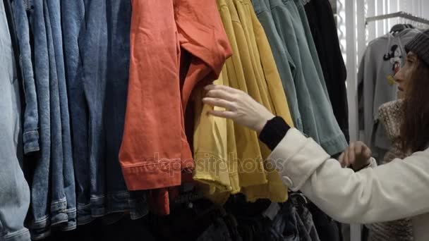 Elinde yeni giysiler, askı askı üzerinde farklı rahat renkli giysiler ile bakarak seçmek genç güzel kadın moda alışveriş merkezinde alışveriş yakın çekim - Video, Çekim