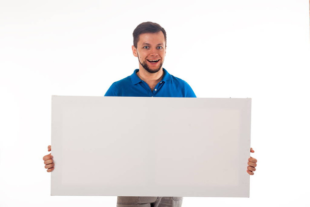 A guy with a beard posing with a white sign. Can be used to place advertising, logo, and other. Dressed in a blue T-shirt and grey pants.  - Photo, image