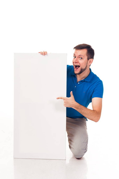 Ένας άντρας με γένια ποζάρει με ένα άσπρο σημάδι. Μπορεί να χρησιμοποιηθεί για να τοποθετήσετε διαφημίσεις, λογότυπο και άλλα. Ντυμένος με ένα μπλε T-shirt και γκρι παντελόνι.  - Φωτογραφία, εικόνα