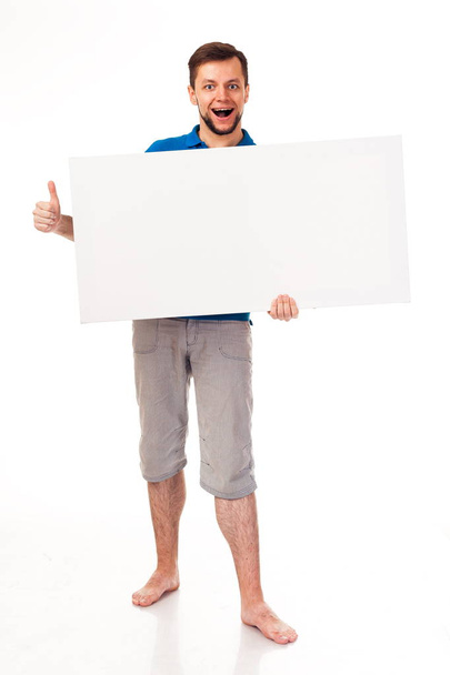 Chlap s plnovousem pózuje s bílou cedulí. Lze použít k umístění reklamy, loga a další. Oblečený v modré tričko a šedé kalhoty.  - Fotografie, Obrázek