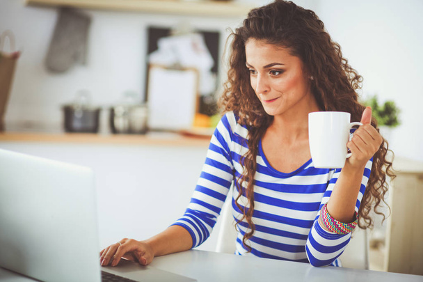 Smiling young woman with coffee cup and laptop in the kitchen at home. Smiling young woman - Foto, Imagen