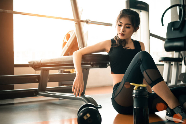 Азиатская молодая женщина отдыхает в фитнес-зале и спортклубе центр с оборудованием и гантели. Концепция тренировок и силовых тренировок. Красота и здоровая тема. Фон гимназии
 - Фото, изображение