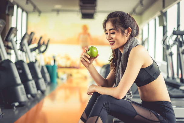 Asian woman holding and looking green apple to eat with sports equipment and treadmill in background. Clean food and Healthy concept. Fitness workout and running theme. - Photo, Image