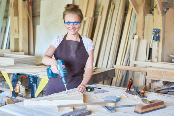 Portrait of cheerful young woman working in carpenters shop drilling holes in piece of wood while making furniture, copy space - Photo, Image