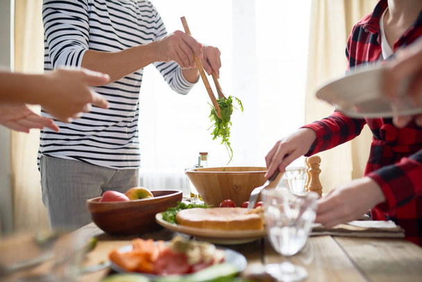 Seitenansicht einer Gruppe von Menschen, die an einem festlichen Esstisch stehen und während der Feierlichkeiten hausgemachtes Essen auf Teller servieren - Foto, Bild