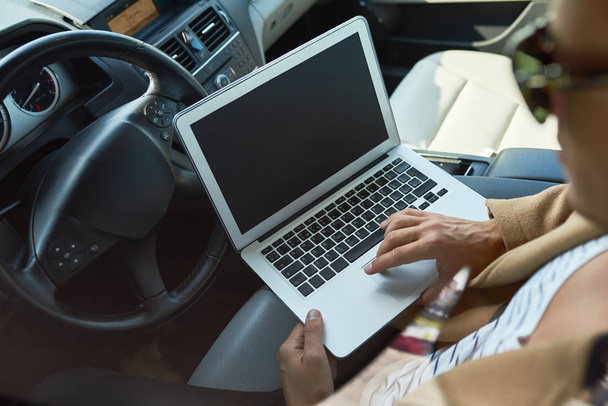 Gros plan à angle élevé de l'homme non reconnaissable réussi à l'aide d'un ordinateur portable assis à l'intérieur d'une voiture coûteuse, espace de copie
 - Photo, image