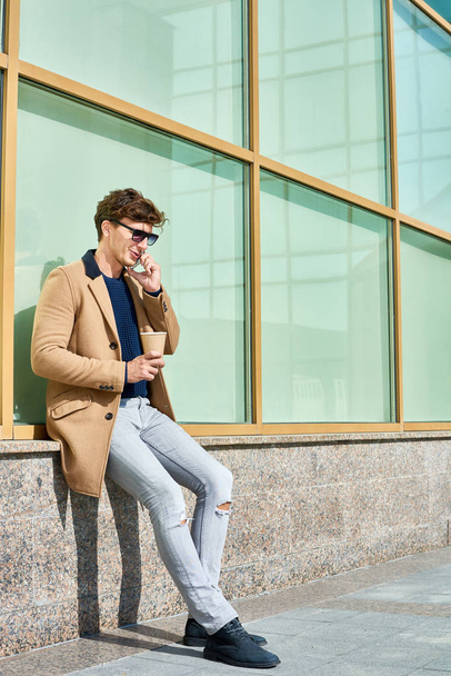 Full length portrait of handsome young man wearing sunglasses speaking by phone while enjoying coffee break outdoors in sunlight, copy space - Photo, Image