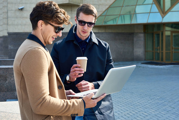 Retrato de dos jóvenes guapos con gafas de sol usando el ordenador portátil y charlando durante la pausa para tomar café al aire libre, espacio para copiar
 - Foto, imagen