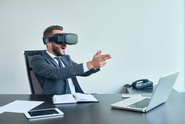 Retrato de hombre de negocios moderno guapo usando gafas VR jugando juego de disparos sentado en el escritorio en la oficina
 - Foto, Imagen