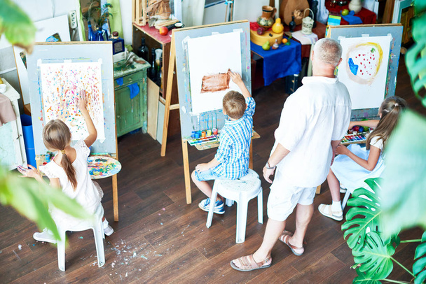 Μεγάλη γωνία πορτρέτο του τρία παιδιά ζωγραφική σε καβαλέτα κατά τη διάρκεια της κατηγορίας τέχνης και δάσκαλος βλέποντας τους στο φιλόξενο στούντιο διακοσμημένα με φυτά - Φωτογραφία, εικόνα