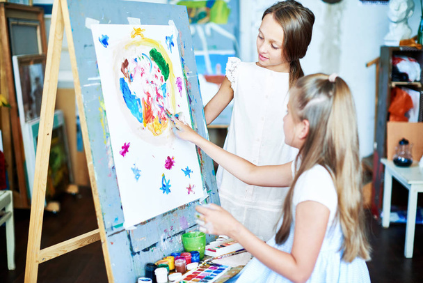 Kaunis pieni tyttö seisoo maalaustelineessä ja piirtää sormin, kun hänen paras ystävänsä tarkkailee hänen toimiaan mielenkiinnolla, tilavan taidestudion sisätilat taustalla
 - Valokuva, kuva