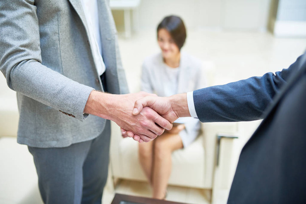 High angle close up of two successful businessmen shaking hands during meeting after closing important deal, young Asian woman in background - Photo, Image