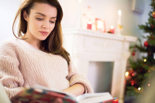 Cintura-up retrato de mulher atraente vestindo suéter sentado no sofá aconchegante, enquanto embrulhado em leitura de livro interessante, interior da sala de estar decorado para a celebração de Natal no fundo
 - Foto, Imagem