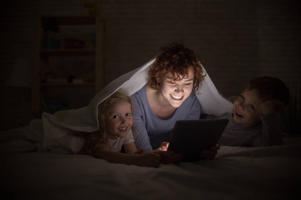 Portrait d'une jeune mère heureuse avec deux enfants lisant des histoires au lit, se cachant sous une couverture et utilisant un livre électronique
 - Photo, image