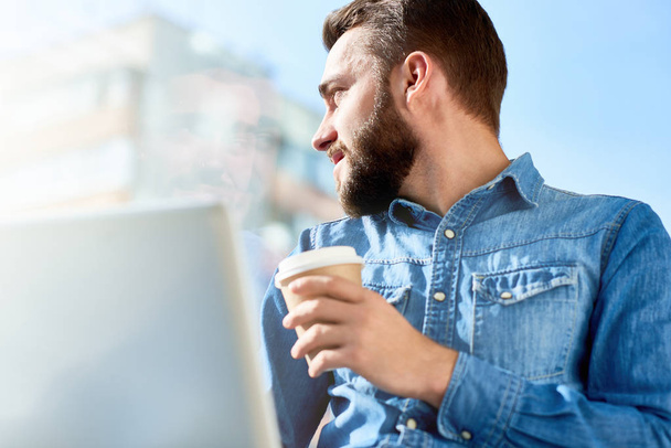 Вид на профіль бородатого молодого менеджера, який дивиться вбік, сидячи на відкритому повітрі і насолоджуючись запашною кавою, розмитим фоном
 - Фото, зображення