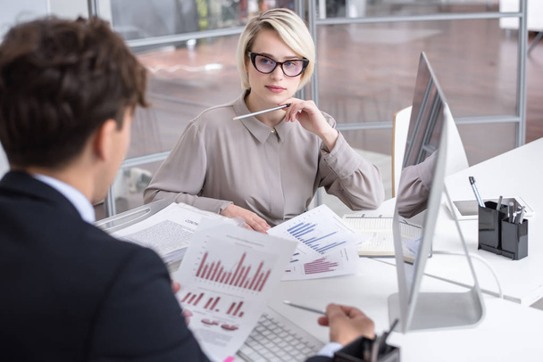 Porträt einer jungen blonden Geschäftsfrau, die während eines Treffens im modernen Büro mit einem Kollegen über Statistiken diskutiert und dabei Diagramme und Diagramme mit Finanzdaten betrachtet - Foto, Bild
