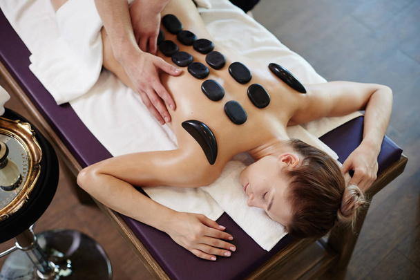 Portrait en angle élevé de jeune femme bénéficiant d'une thérapie de pierre allongée sur une table de massage au centre SPA
 - Photo, image