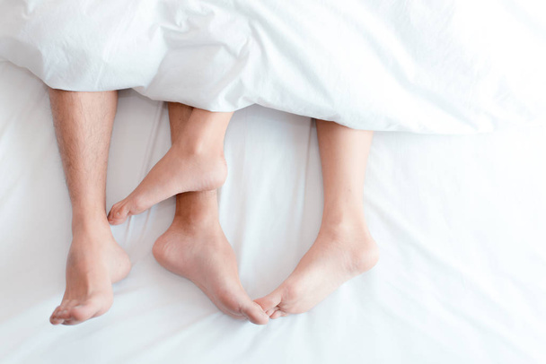 Closeup feet of couple on the bed. Man and woman lovers make love under the blanket or bed sheet. Sex on vacation theme. Valentine and Honeymoon concept. - Photo, Image