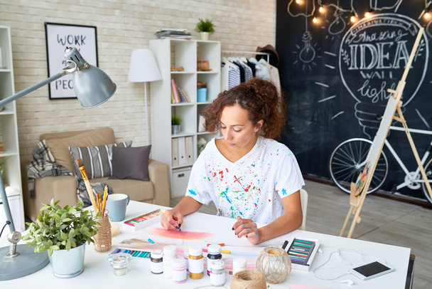 Портрет творчої молодої жінки, що малює пастельну картину, що сидить за столом в сучасній квартирі
 - Фото, зображення