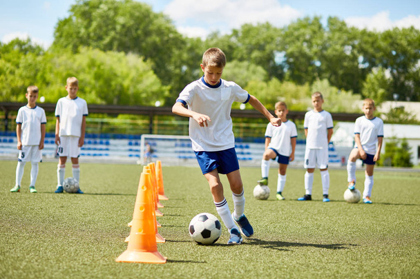 Retrato de niño en el equipo de fútbol junior liderando la pelota entre los conos durante la práctica en el campo en el día soleado
 - Foto, imagen
