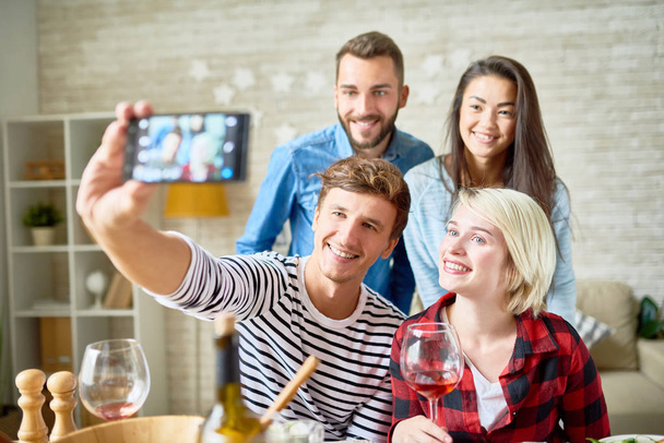 Portrait of four happy friends taking selfie at dinner table while celebrating holiday together at home - Foto, Bild