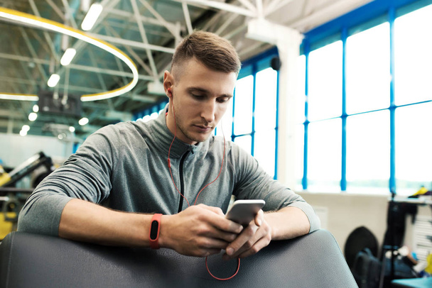 Portrait of handsome young man using smartphone listening to music in modern gym taking break from workout - Foto, afbeelding