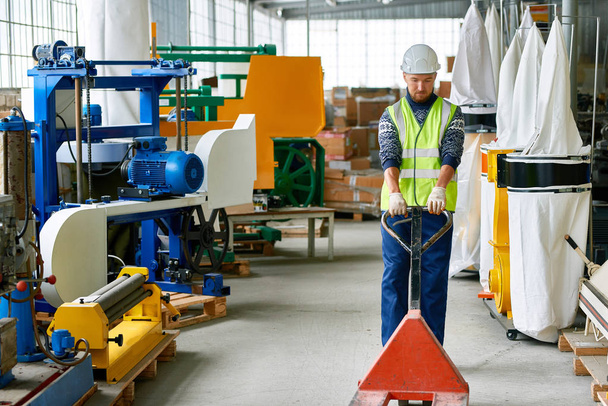 Retrato de comprimento total do trabalhador usando chapéu duro puxando carrinho vazio na oficina industrial da fábrica moderna, espaço de cópia
 - Foto, Imagem